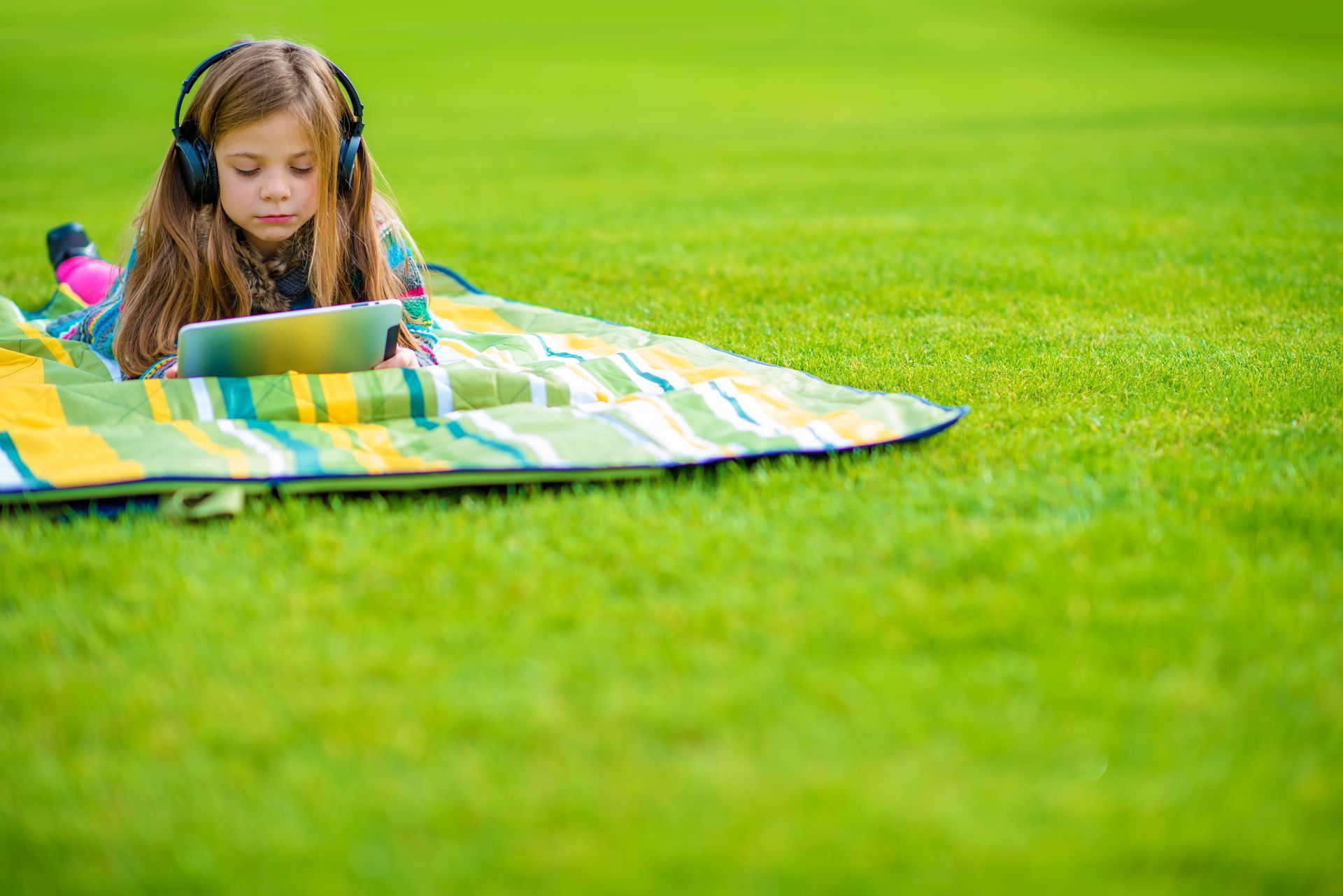 Girl wearing headphones, lying on a blanket on the grass, looking at a tablet.