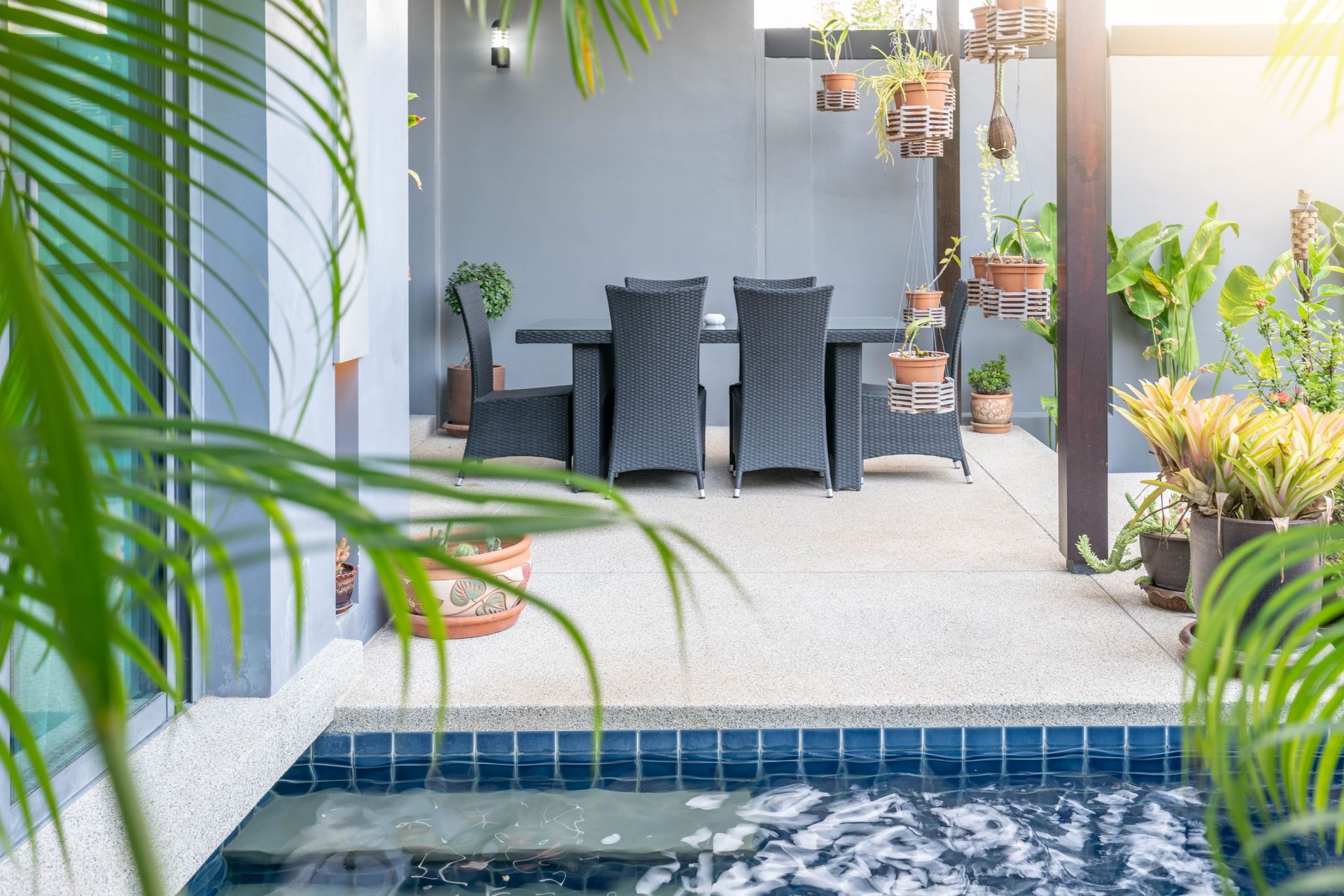 Patio with wicker furniture near a pool. Plants, gray walls, and sunlight are visible.