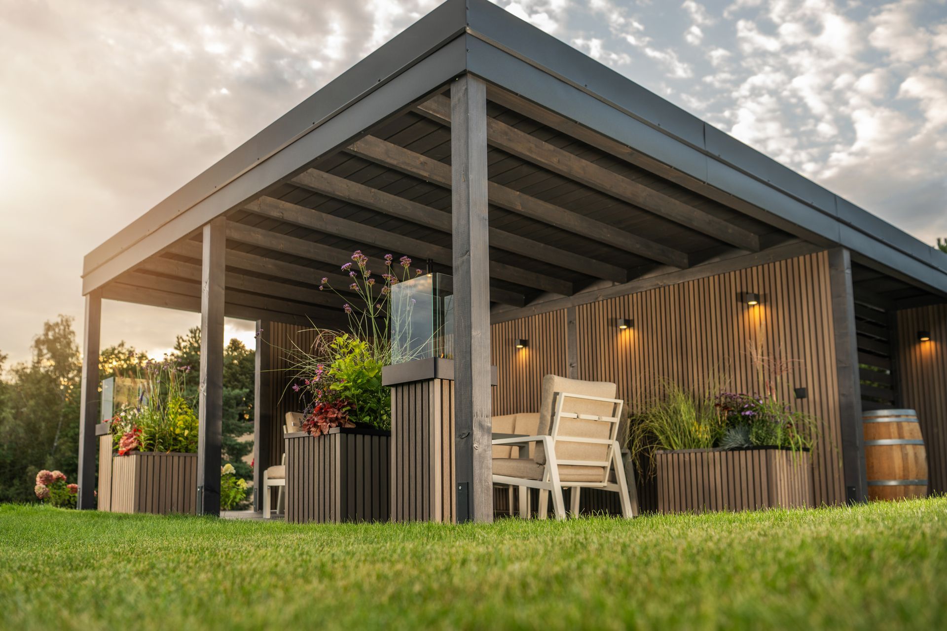 Outdoor pergola with seating, plants, and wooden accents, set against a grassy lawn and cloudy sky.
