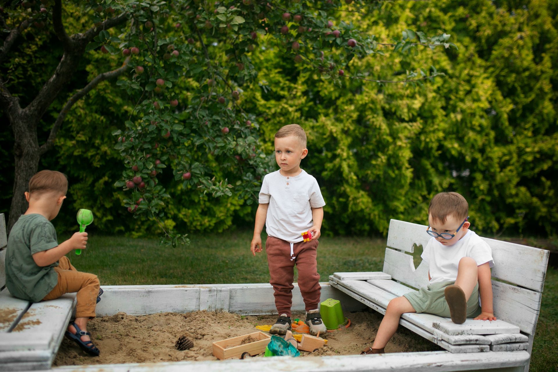 Three children playing in a sandbox with toys, sitting on a bench. Green and brown colors.