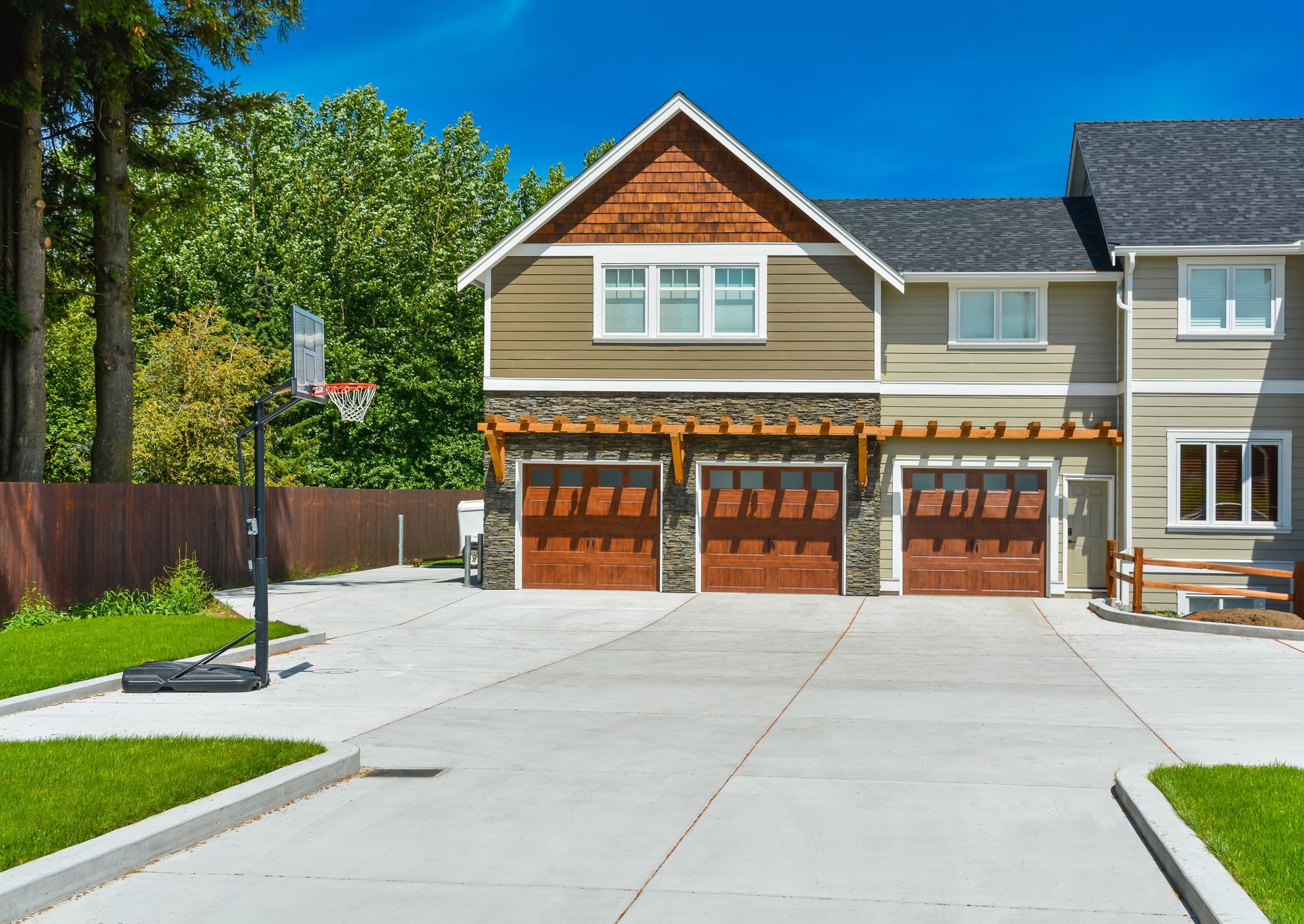Three-car garage with brown doors and pergola, driveway, and basketball hoop in front of a house.
