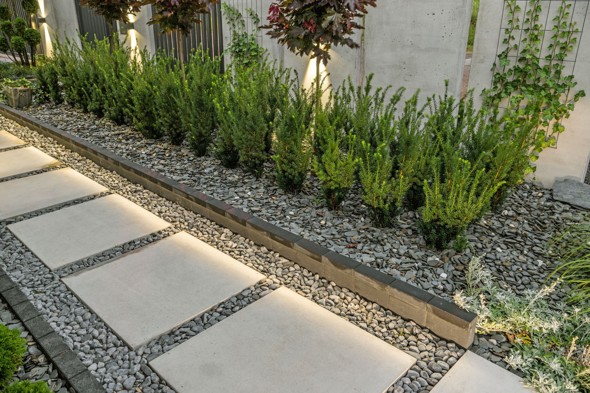 Stone path with illuminated edges next to a bed of green shrubs and pebbles.