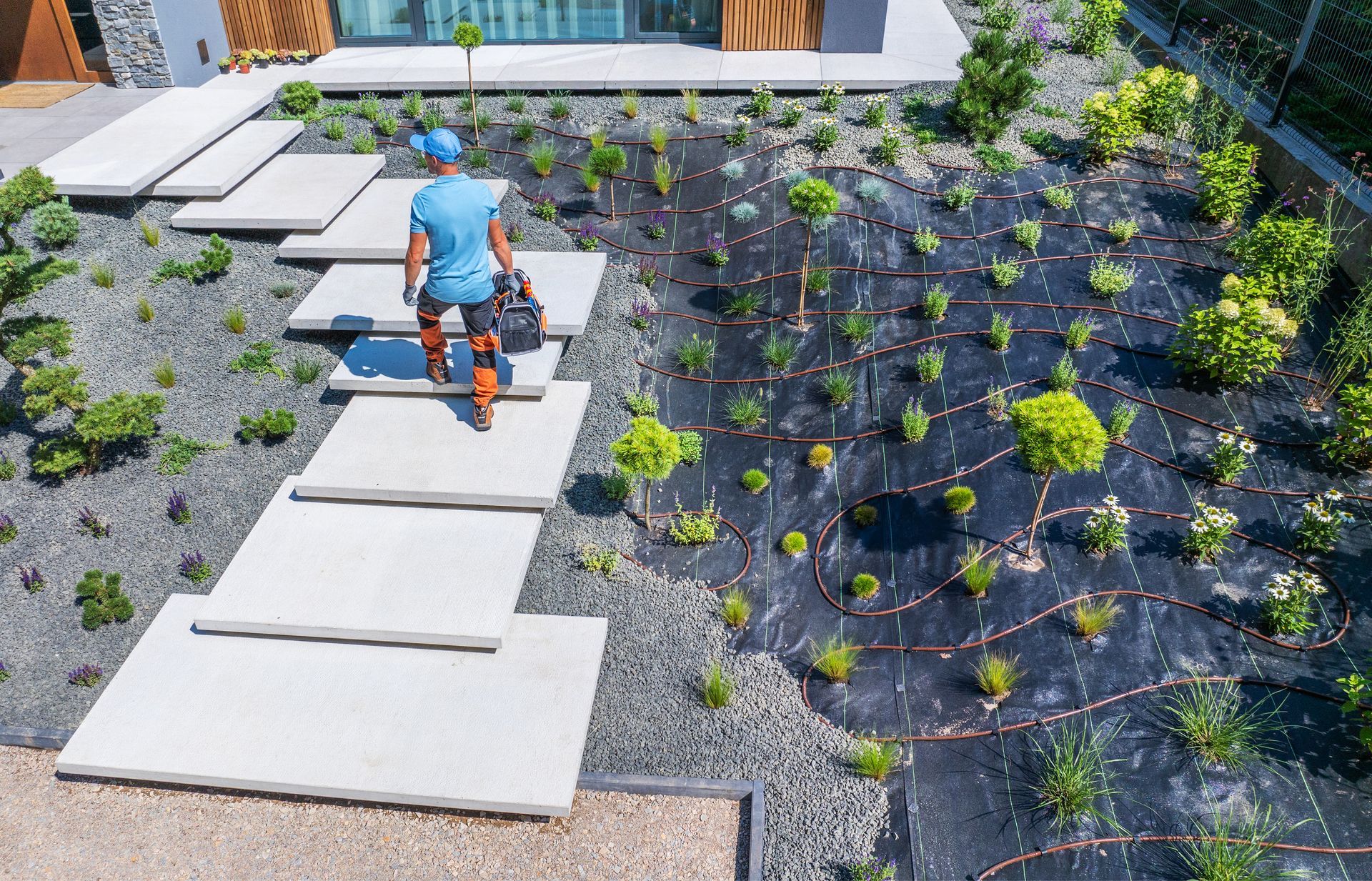 Man walking up concrete steps with gardening equipment; landscaped garden with plants.