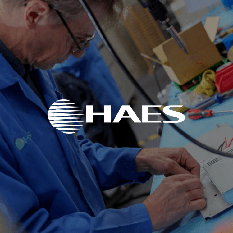 A man in a blue coat is working on an electronic device with the haes logo behind him