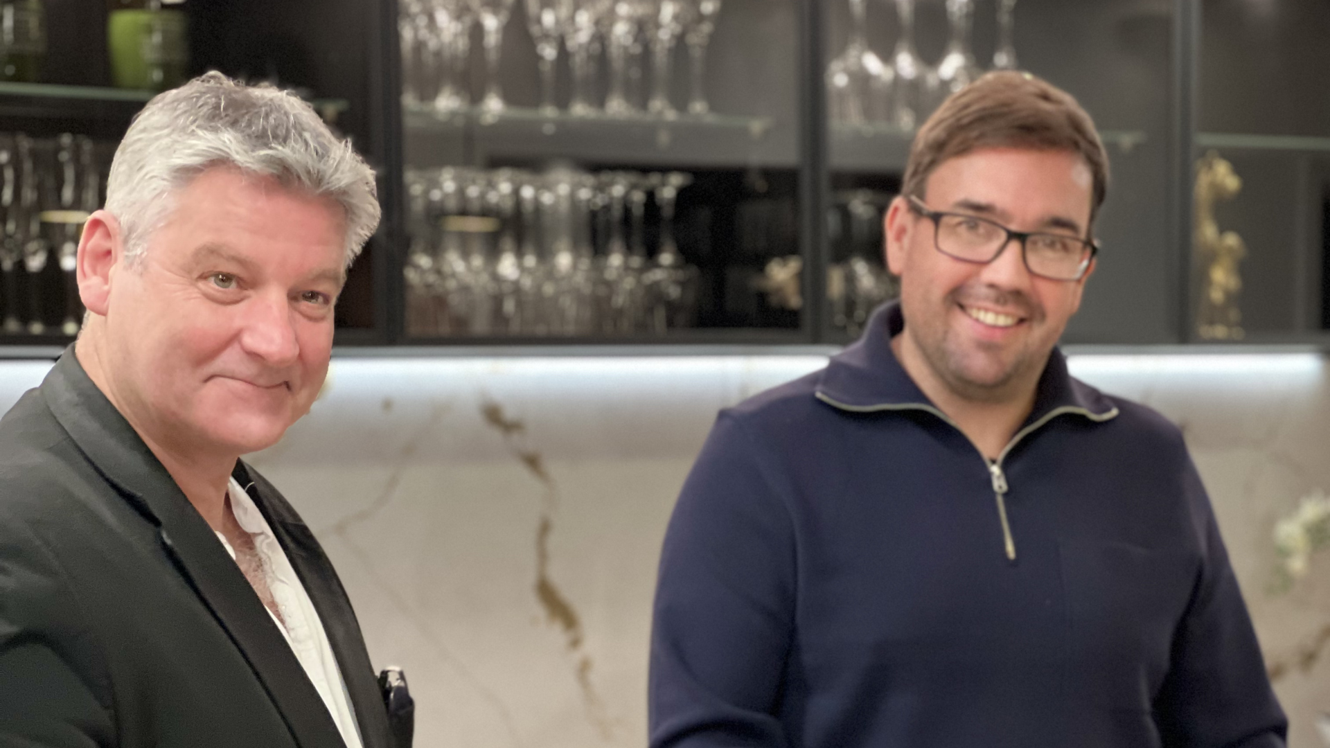 Someone holds a phone to record two people in a kitchen standing by a marble island with an open magazine.