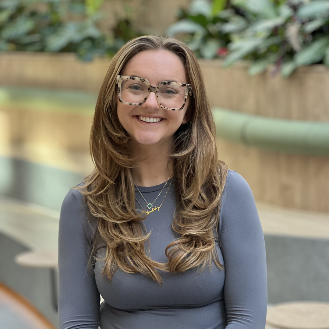 A smiling person with long blonde hair wearing a black polka-dot blouse, standing in an office with a green wall.