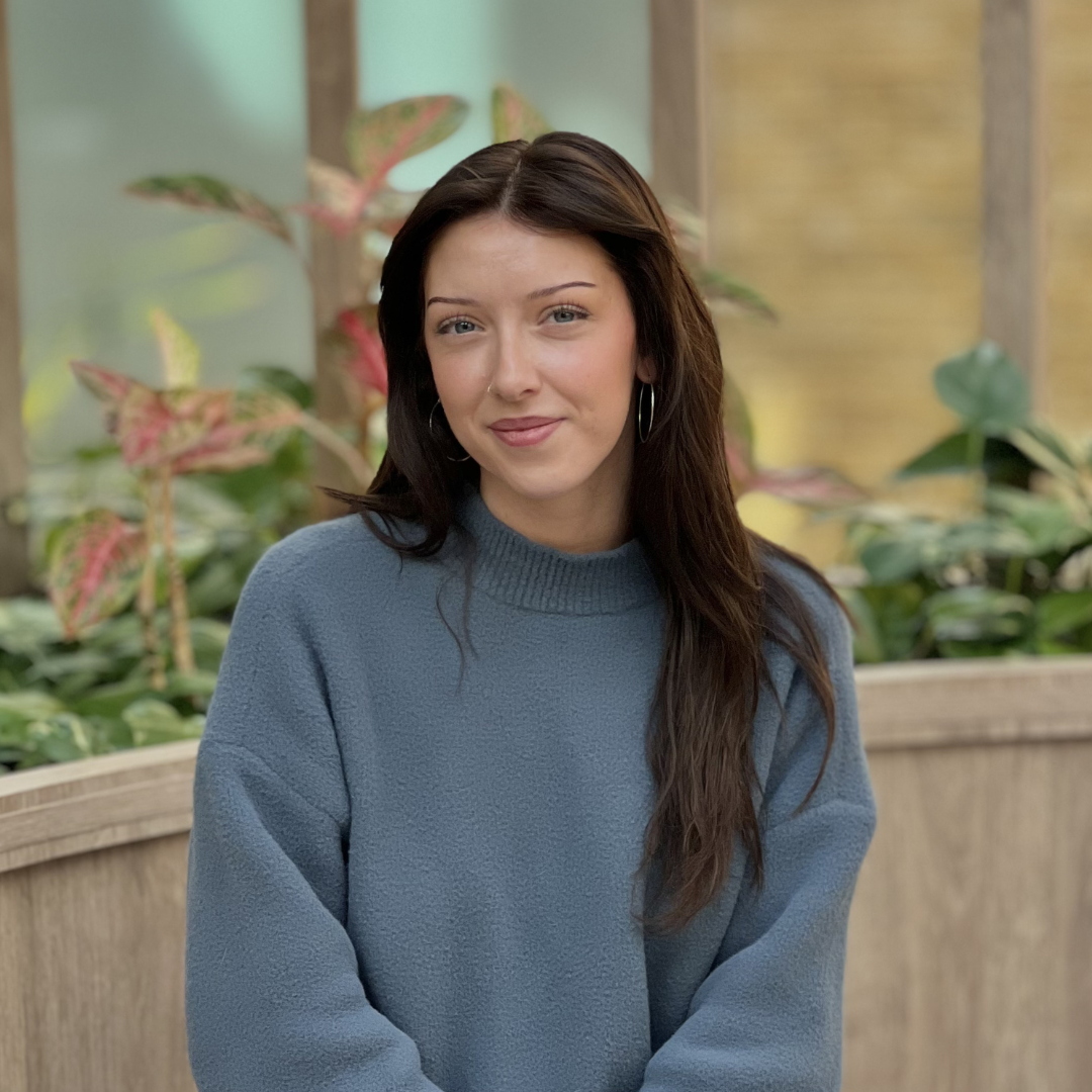 A person with shoulder-length brown hair and a neutral expression wears a grey sweater and a gold layered necklace.