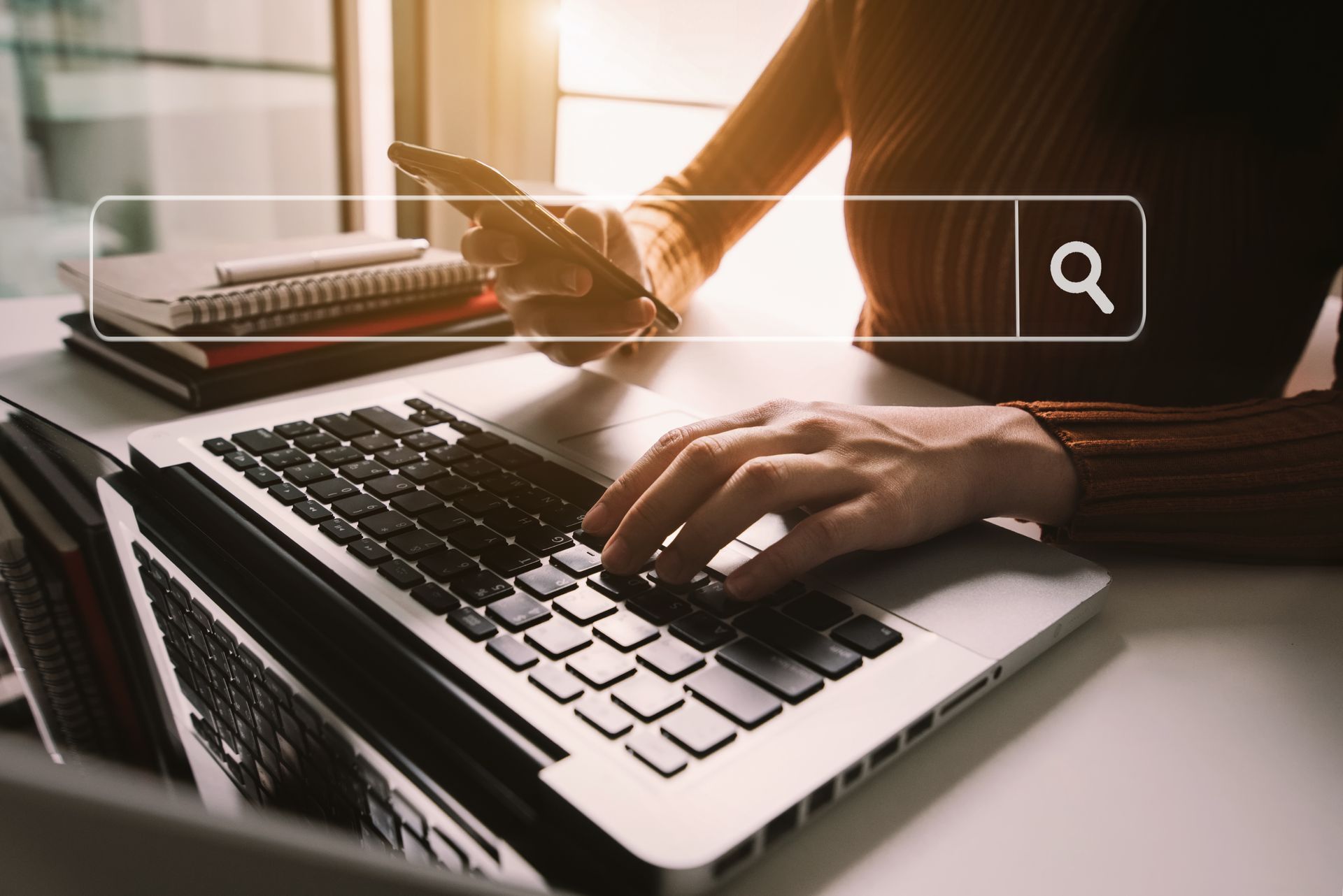 Person typing on laptop, holding phone, search bar overlay.