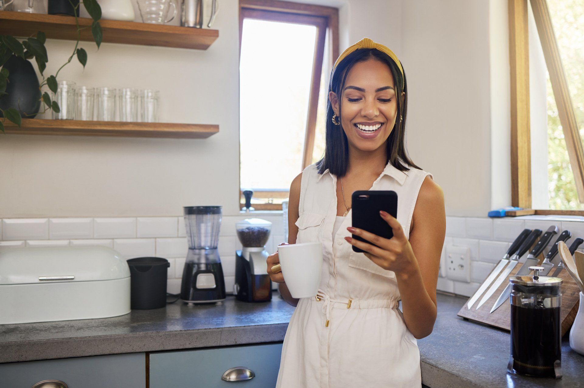 woman standing in a kitchen smiling at her phone screen & holding a mug