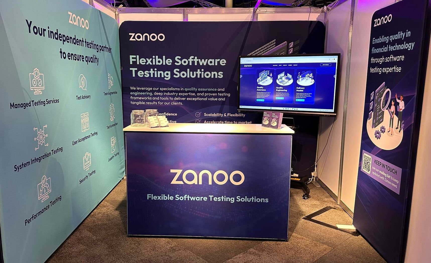 Trade show booth for Zanoo software testing. Dark blue and light teal with screen display and table.