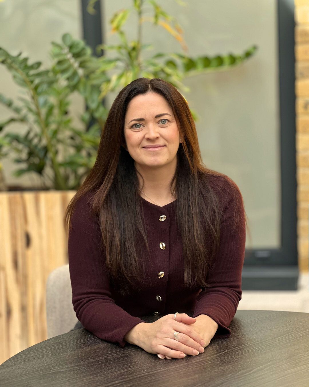 A person with long brown hair smiling at the camera, wearing a black shirt with white polka dots against a gray wall.