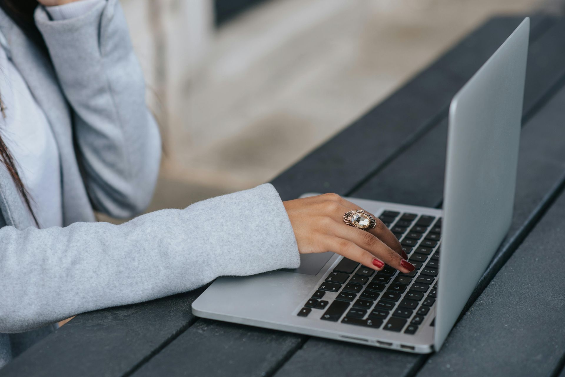 Woman's hand typing on a laptop, wearing a ring, on a dark wooden table outside.