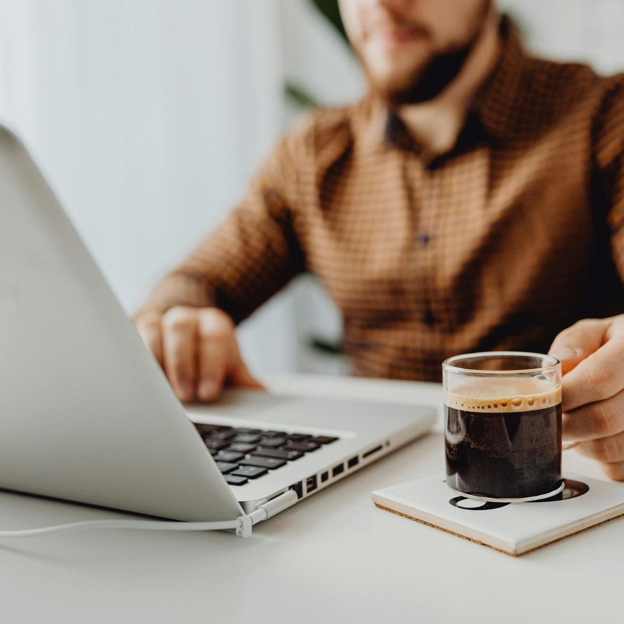 A person in a brown patterned shirt uses a laptop at a white desk while holding a glass mug of black coffee.