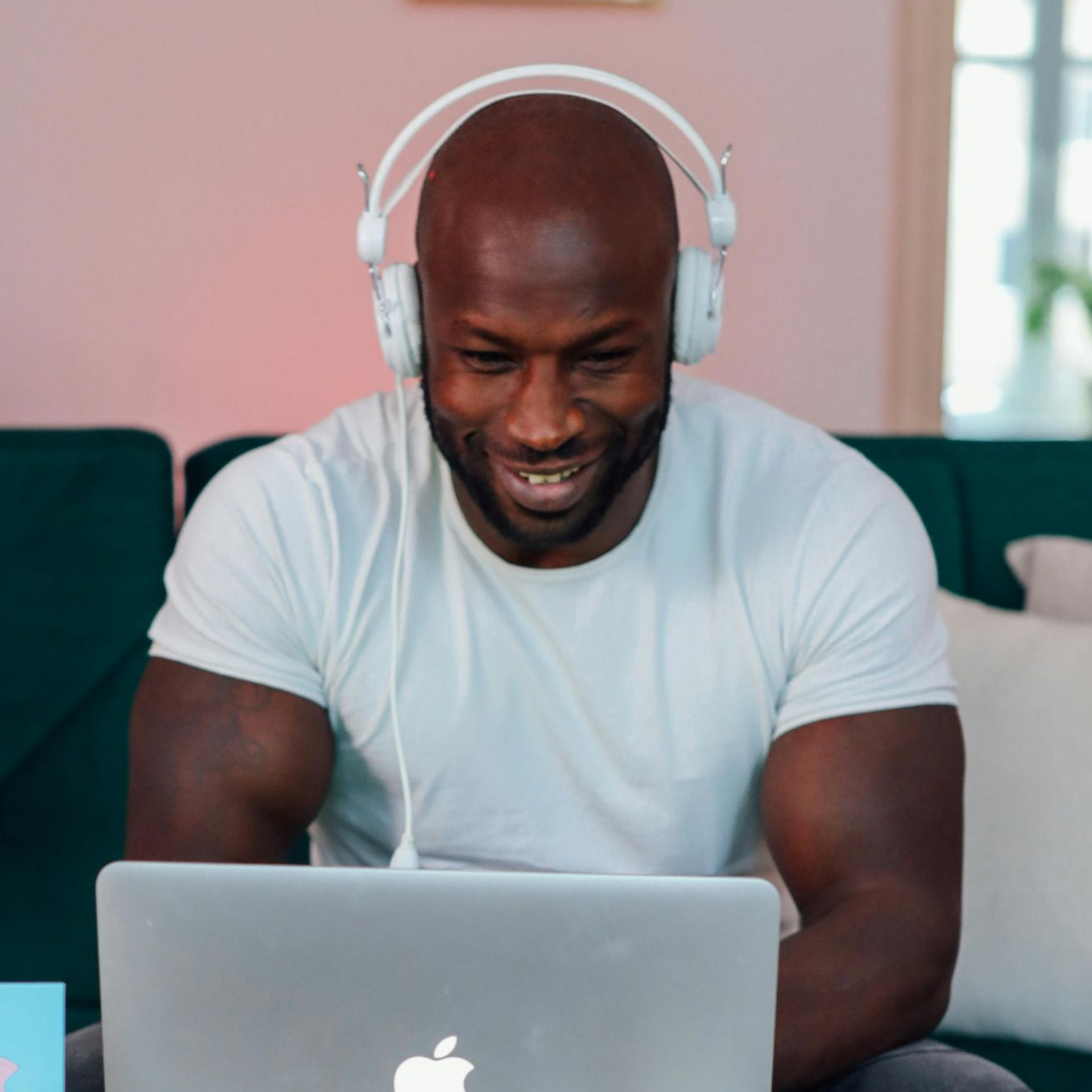 A person with a shaved head wears white headphones and a white t-shirt while smiling at a laptop in a bright room.