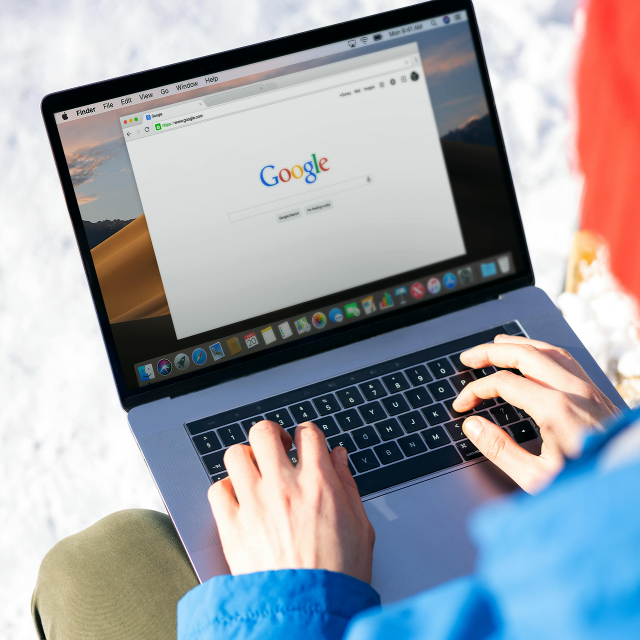Hands type on a silver laptop showing the Google homepage, with a person in a blue jacket outdoors in a snowy setting.