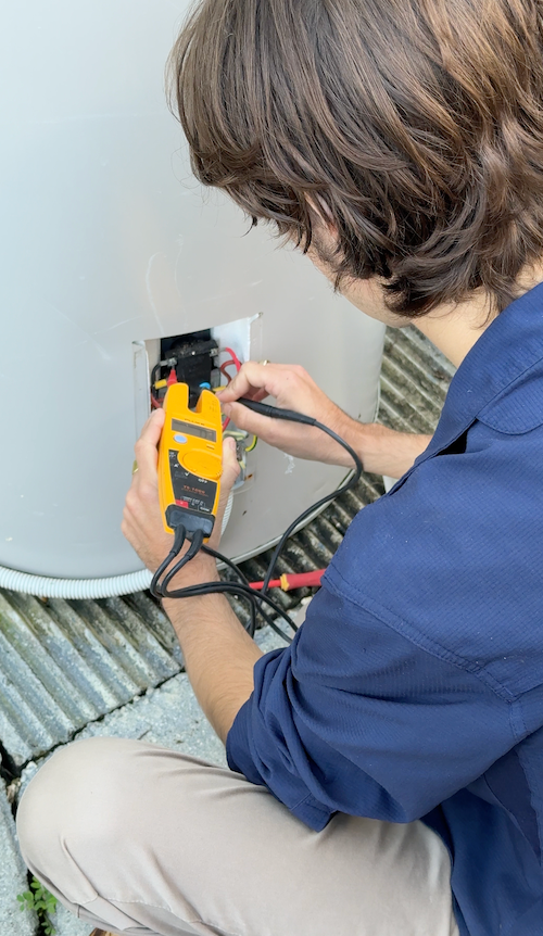 Technician using a clamp meter to inspect HVAC wiring beside a large unit.