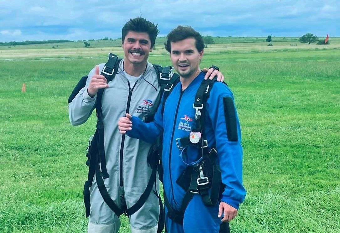 Two men in parachute suits are posing for a picture in a field.
