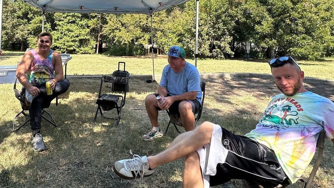 A group of men are sitting in chairs under an umbrella in a park.