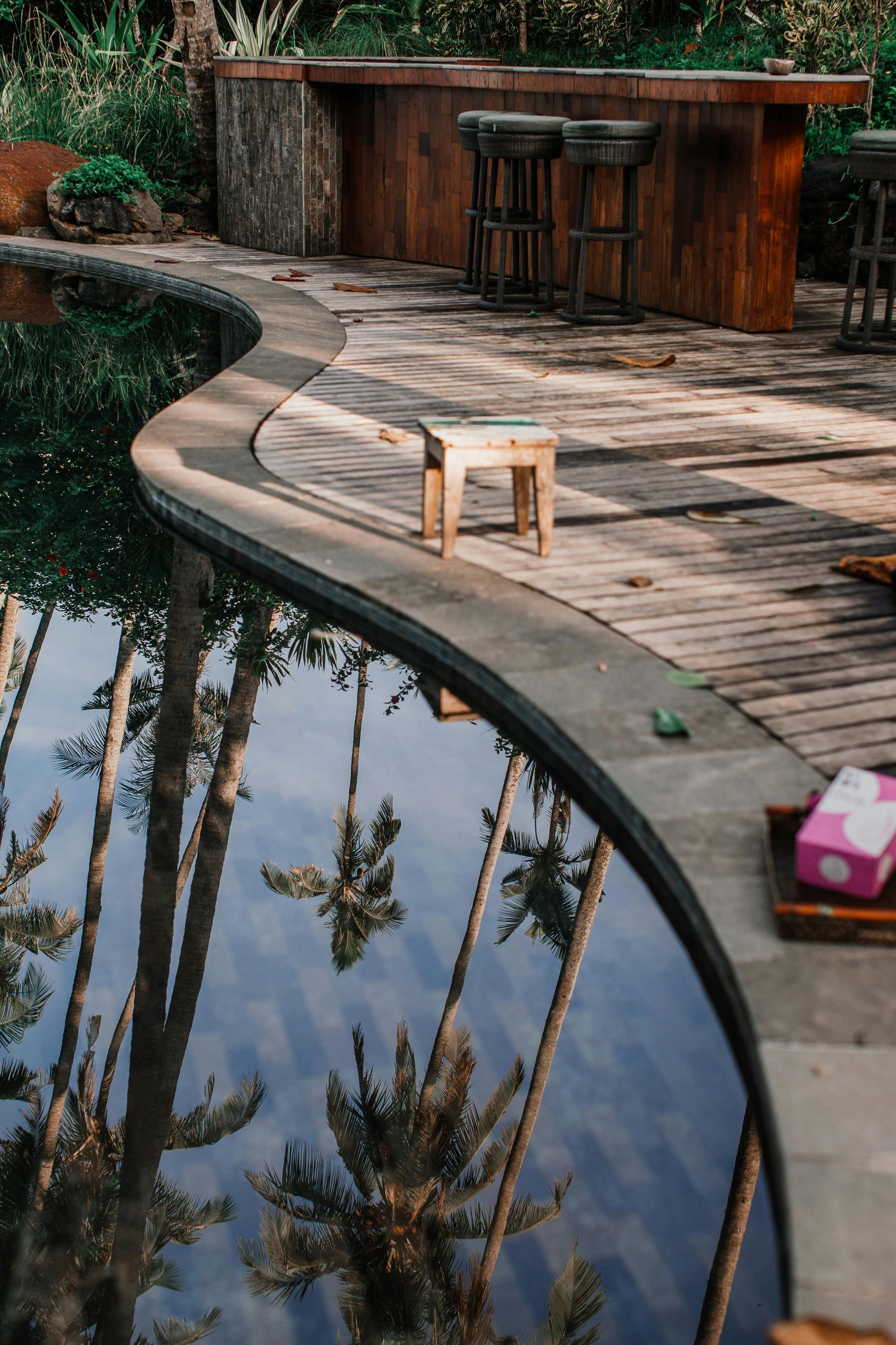 A wooden deck next to a swimming pool with palm trees reflected in the water.