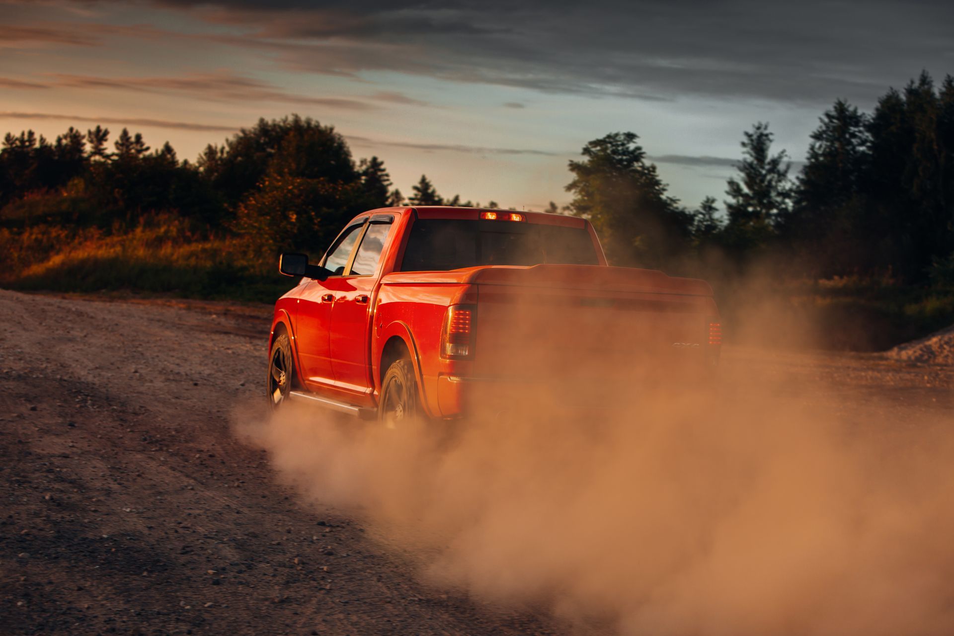 Red pickup truck kicking up dust on a dirt road at sunset.