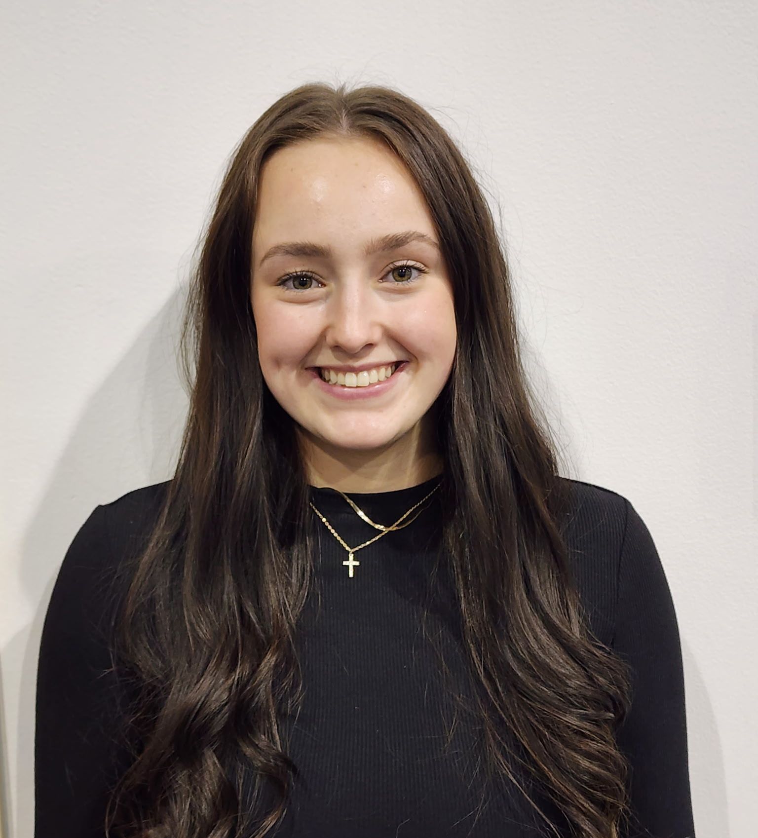 A young woman with long brown hair smiles at the camera. She wears a black top and gold necklace, against a white backdrop.