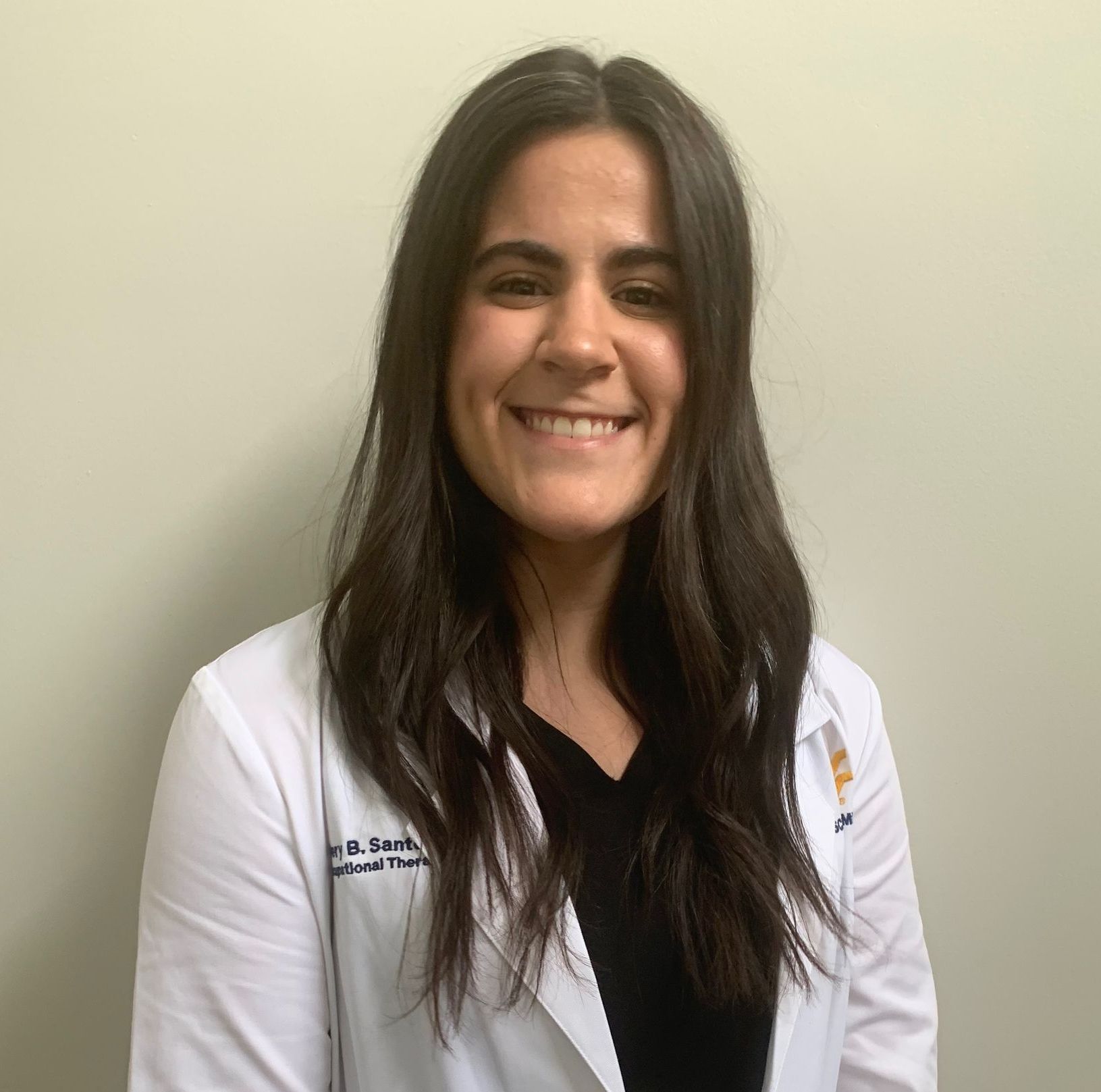 Woman with dark hair, wearing a white coat over a black top, smiles at the camera in front of a neutral-colored wall.