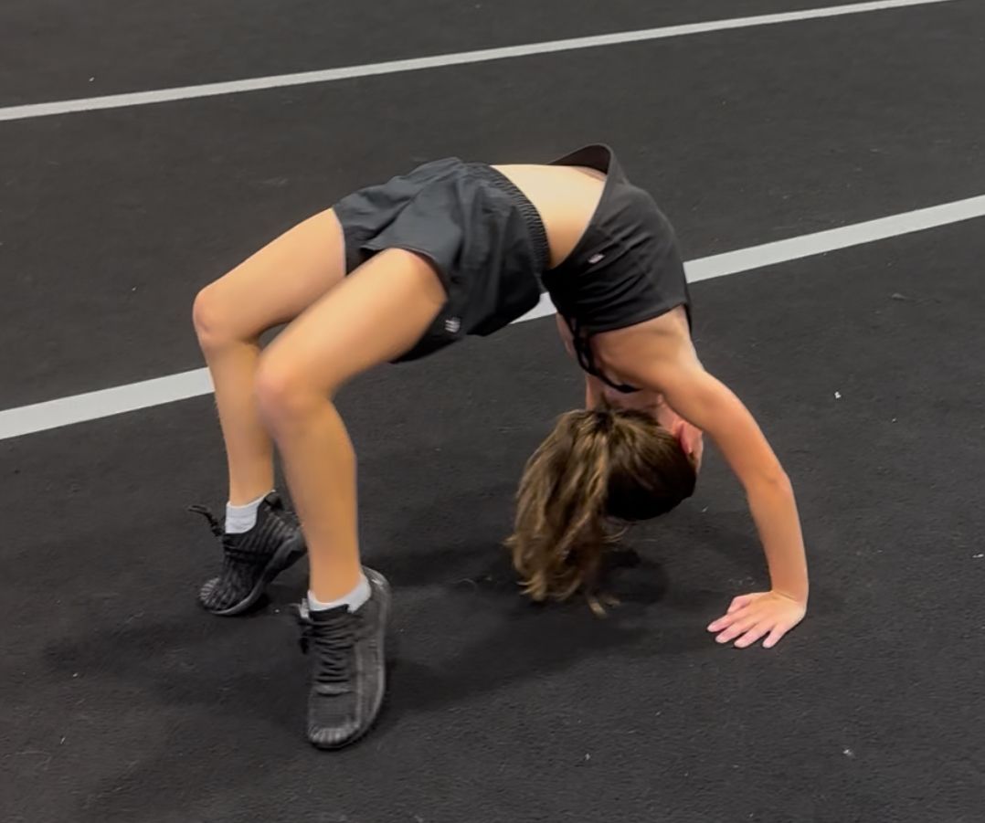 Two young cheerleaders in red and black uniforms doing cartwheels on a blue mat. They have a focused expression.