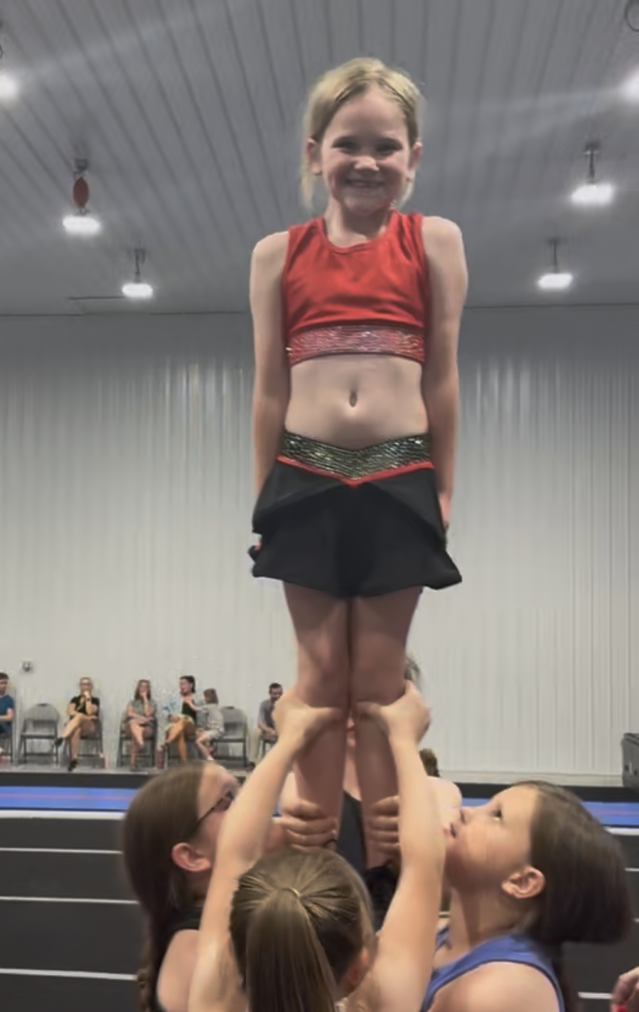 Young girl smiles while being lifted in a cheerleading stunt, wearing a red top and black skirt, inside a gym.