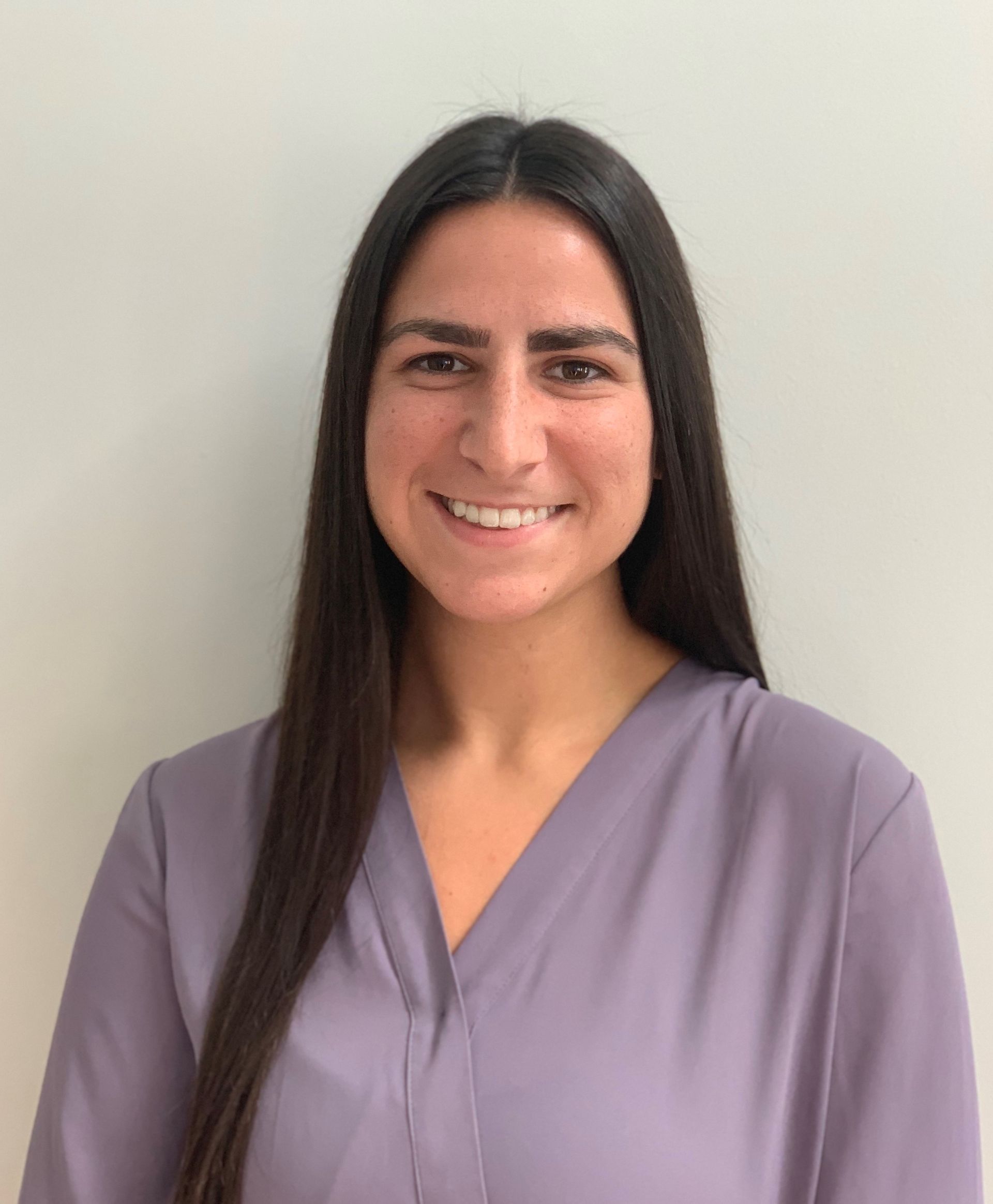 Woman with long, dark hair smiles, wearing a light purple blouse, against a plain white wall.