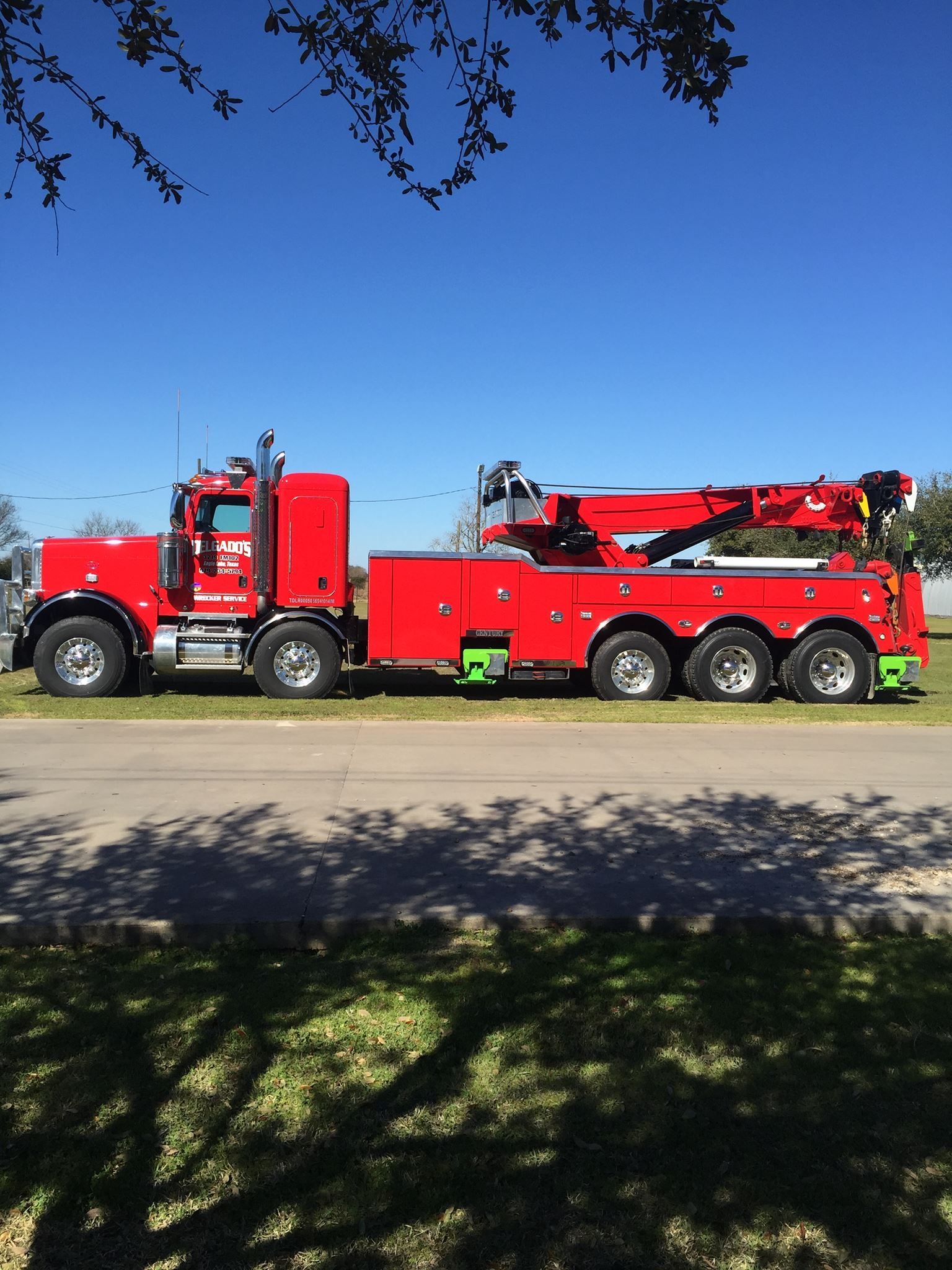 A bright red, multi-axle heavy-duty tow truck parked on a grass verge next to a paved road under a clear blue sky.