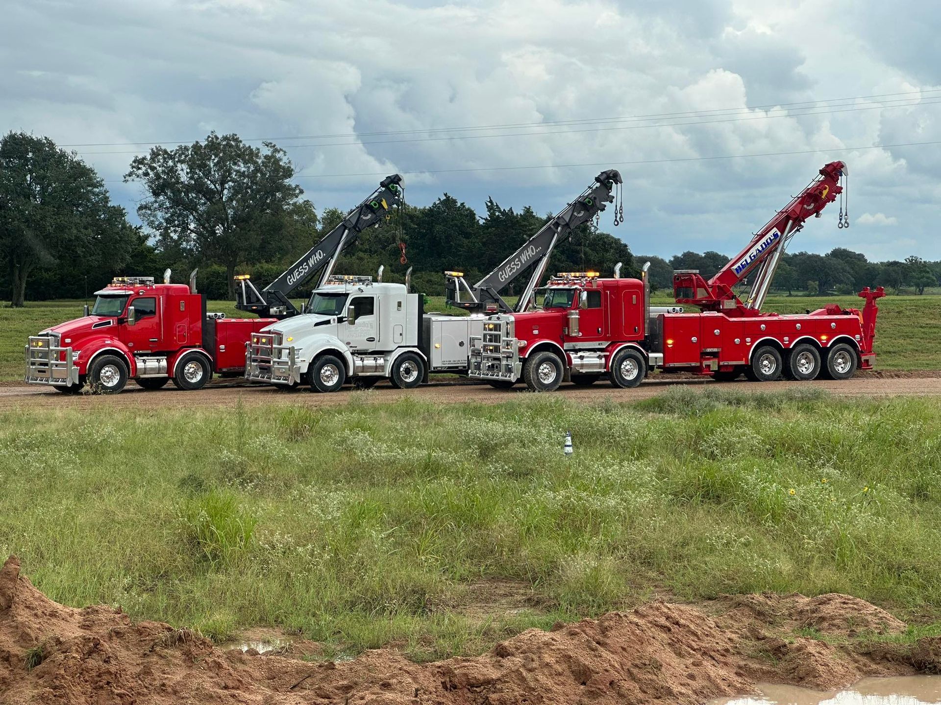 Three large tow trucks—red, white, and red—parked in a line on a grassy field under a cloudy sky.