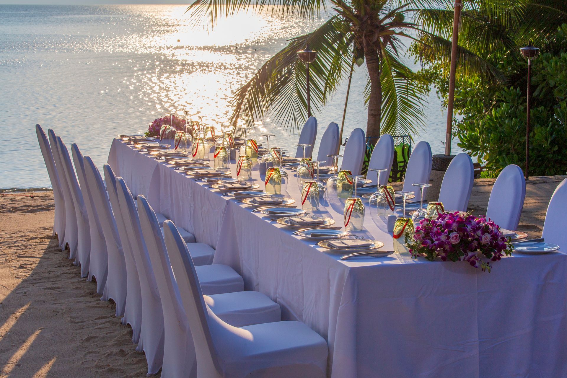 Lungo tavolo apparecchiato per un pasto su una spiaggia assolata, con tovaglie bianche e composizioni floreali.