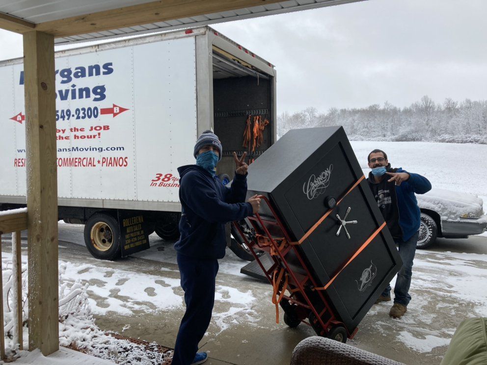 Two men are carrying a large safe on a dolly in front of a moving truck.