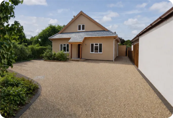 A beige house with a gravel driveway, a small front porch, and a white side wall under a partly cloudy sky.