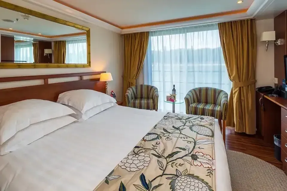 Hotel room with a bed, two chairs, and a large window overlooking water.