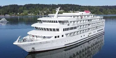 White cruise ship on calm blue water, trees and buildings in the background.