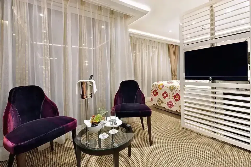A modern hotel room with purple chairs, a coffee table, and a TV behind a slat wall.