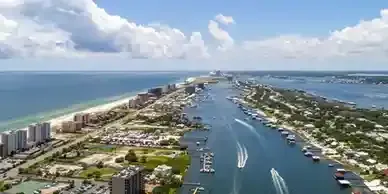 Aerial view of a coastal city with buildings, a waterway, boats, and sandy beaches under a blue sky with clouds.