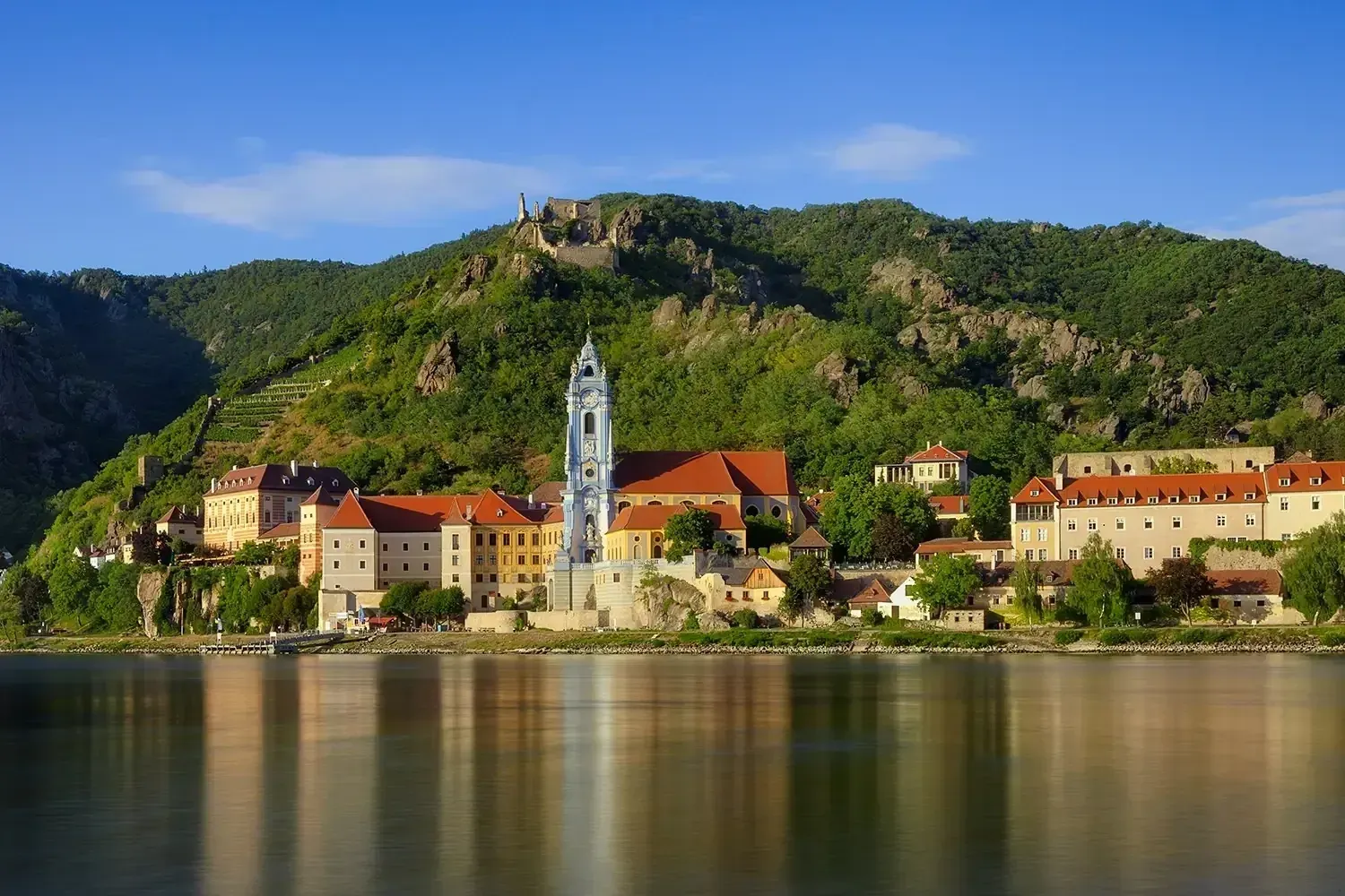 Town of Dürnstein, Austria, reflected in the Danube River. Buildings with red roofs and a prominent church tower are set against a forested hillside.
