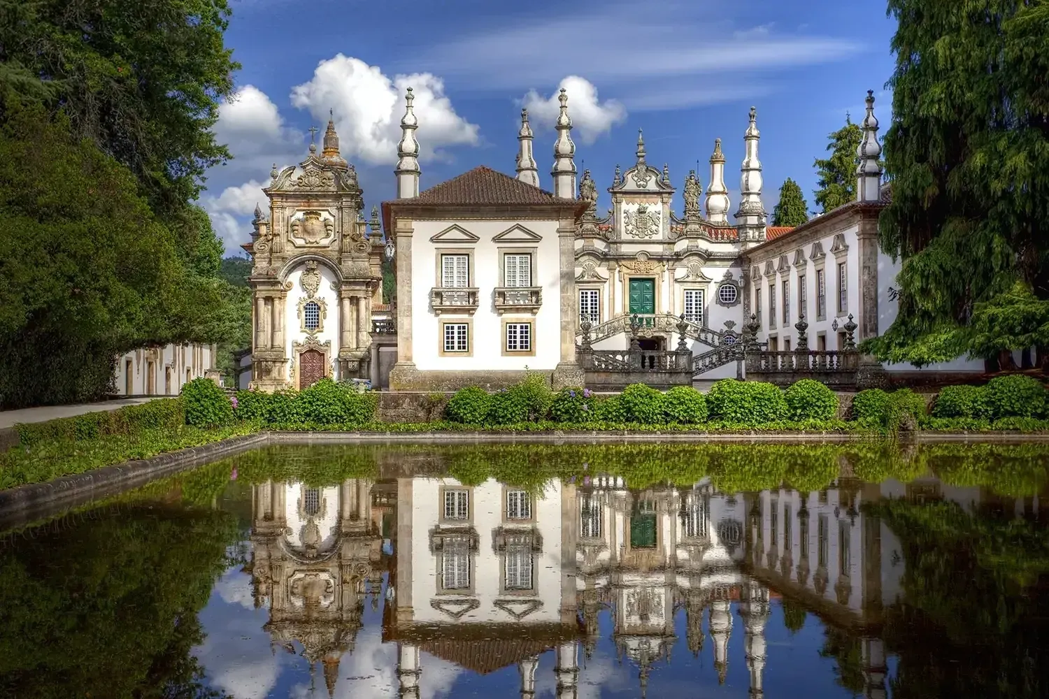Baroque palace, Portugal, reflected in a pond; white facade, ornate details, blue sky, green trees.