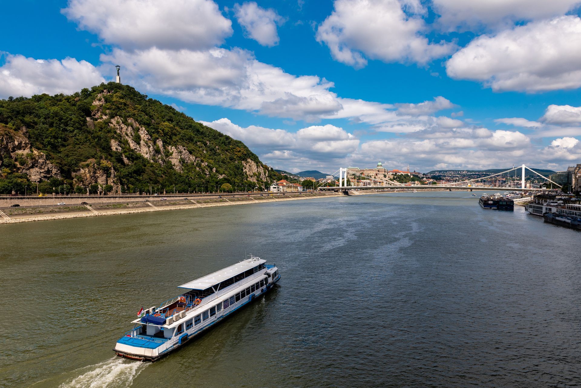 A boat sails on a blue river, under a bridge, between buildings and green hills, under a bright sky.