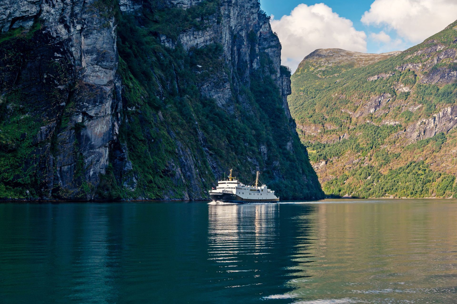 A ferry sails between tall, rocky cliffs on a serene blue fjord, with a mountain and blue sky in the background.