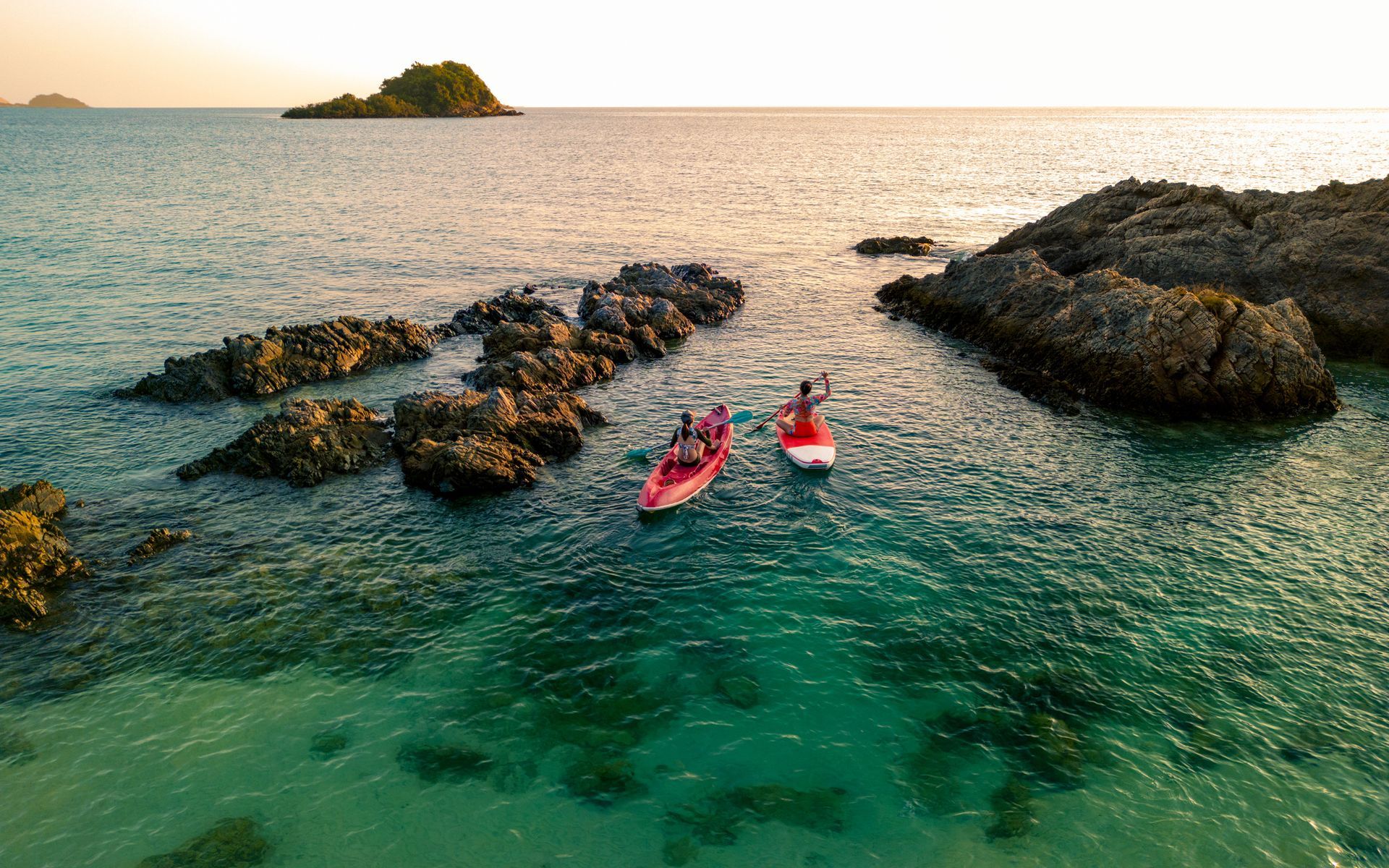 Two people paddleboarding near rocks in clear turquoise water; island in background.