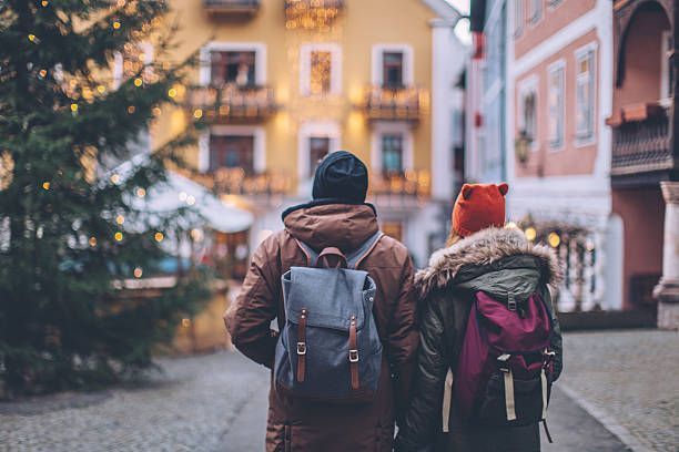 Couple with backpacks walking in a European town; cobblestone street, festive decorations, buildings in soft colors.