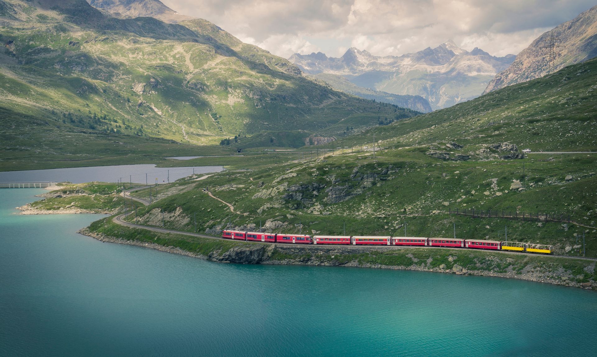 Red train travels along a lake, surrounded by mountains and lush green landscape.