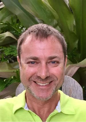 Man with short hair smiling outdoors, wearing a light green shirt; greenery in the background.