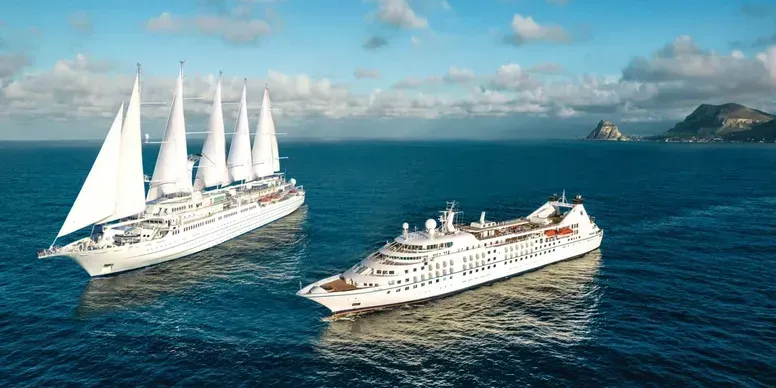 Two white cruise ships are sailing on a blue ocean, with a hilly landmass in the background.