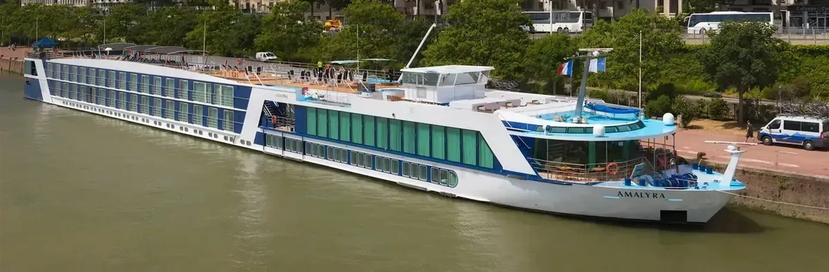 River cruise ship docked along a river with trees in the background. The boat is white and blue.