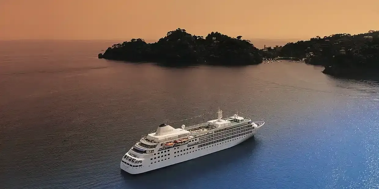 Cruise ship in a bay with dark water and an orange sky, surrounded by green, hilly land.