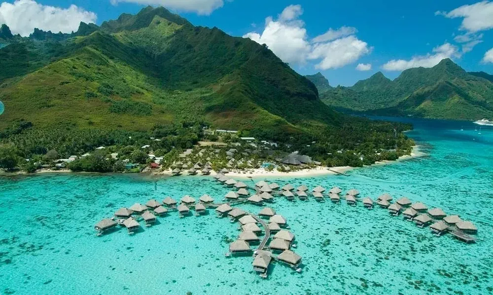 Overwater bungalows in turquoise waters with a lush green mountain backdrop.